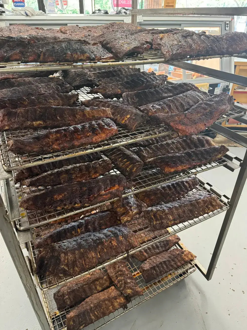 Multiple racks filled with smoked ribs and other cured meats displayed indoors on metal wire shelves.