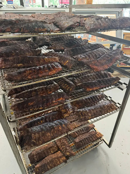 Multiple racks filled with smoked ribs and other cured meats displayed indoors on metal wire shelves.